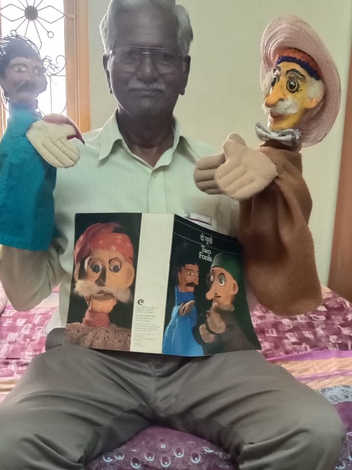 Dr. N. Dhanaraj seated with a glove puppet on each hand, holding the CCRT 'Two Fools' booklet